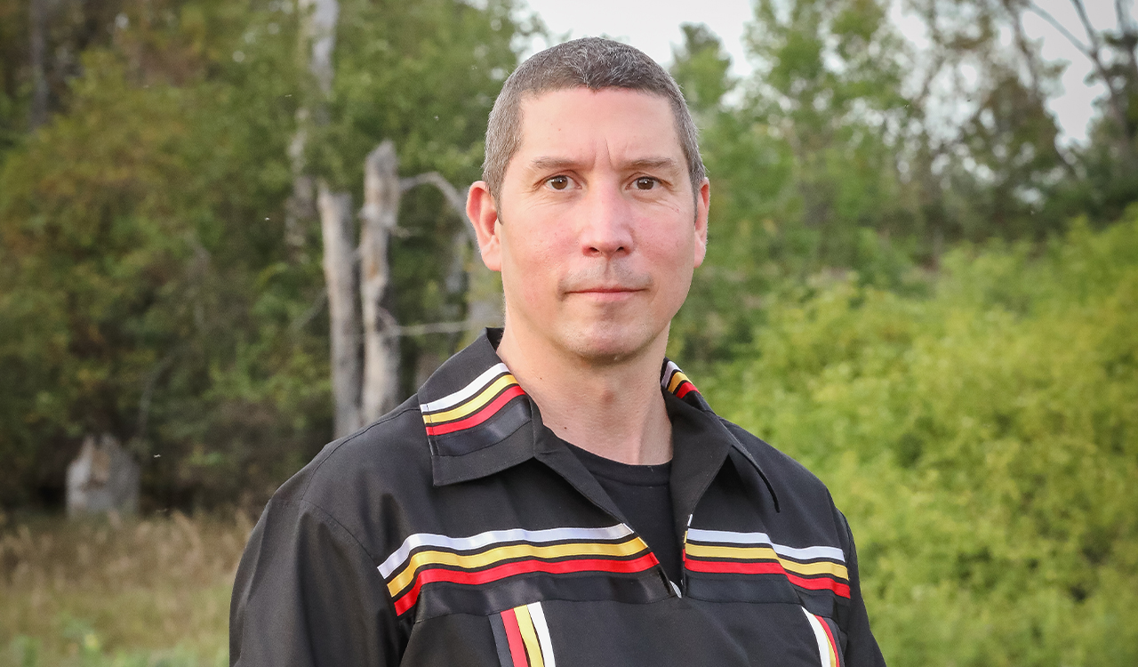 Picture of Marc Forgette, owner of Makatew Workshops, dressed in a black shirt with red and yellow accents, standing in front of trees