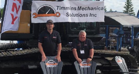 Two Timmins Mechanical Solutions employees  posing in front of mechanical equipment with a company banner and chairs bearing the company logo.