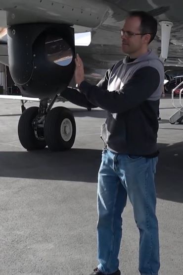A person in civilian clothing adjusts a sphere with a reflective lens, which is mounted under an airplane.